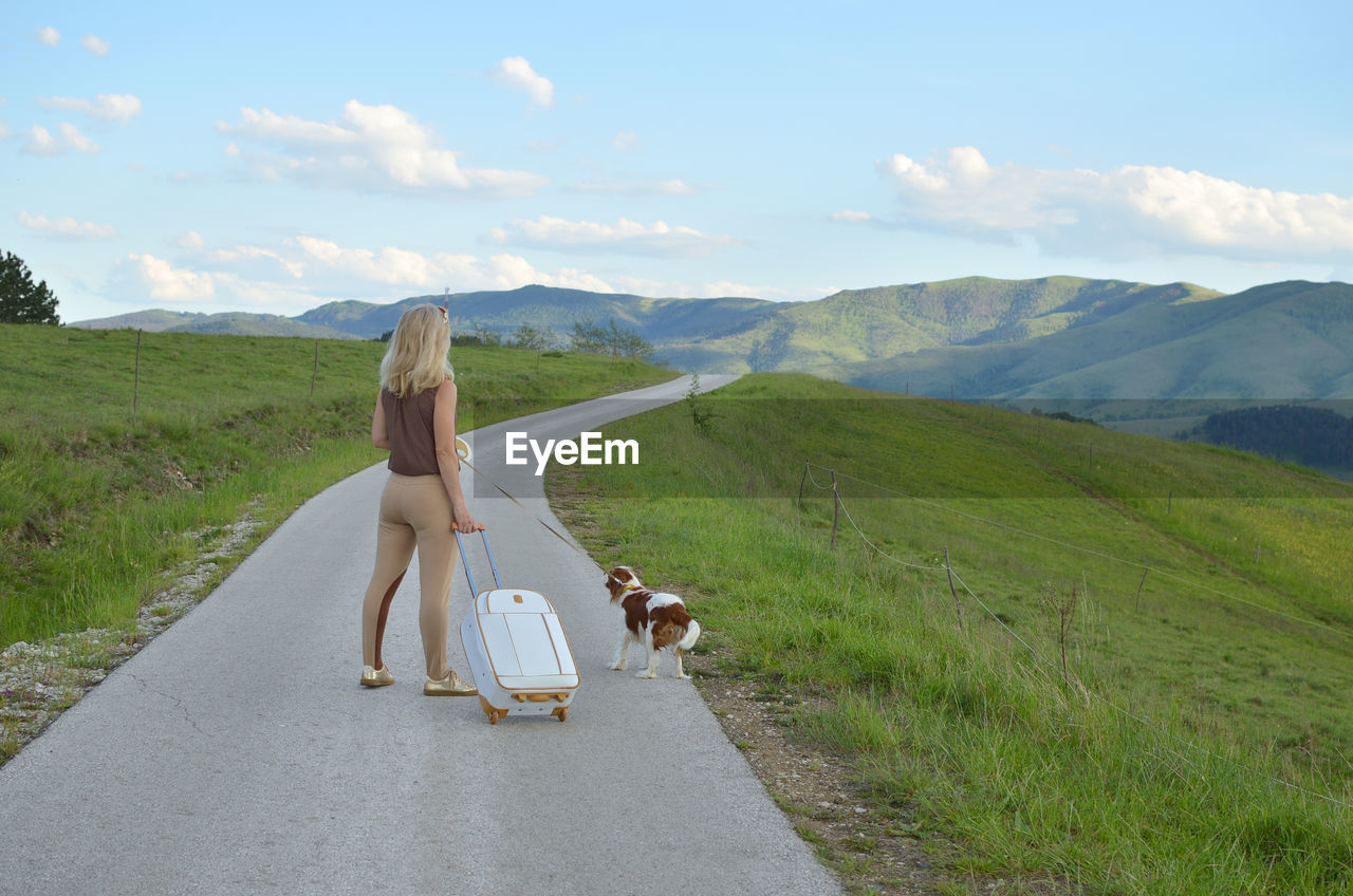 Woman and her dog fellow traveler with suitcase are standing in the middle of a mountain road