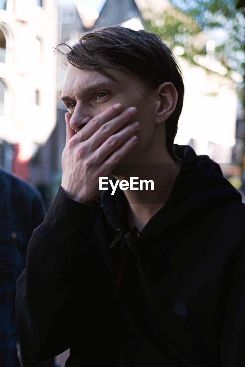 Young man looking away while covering mouth in city