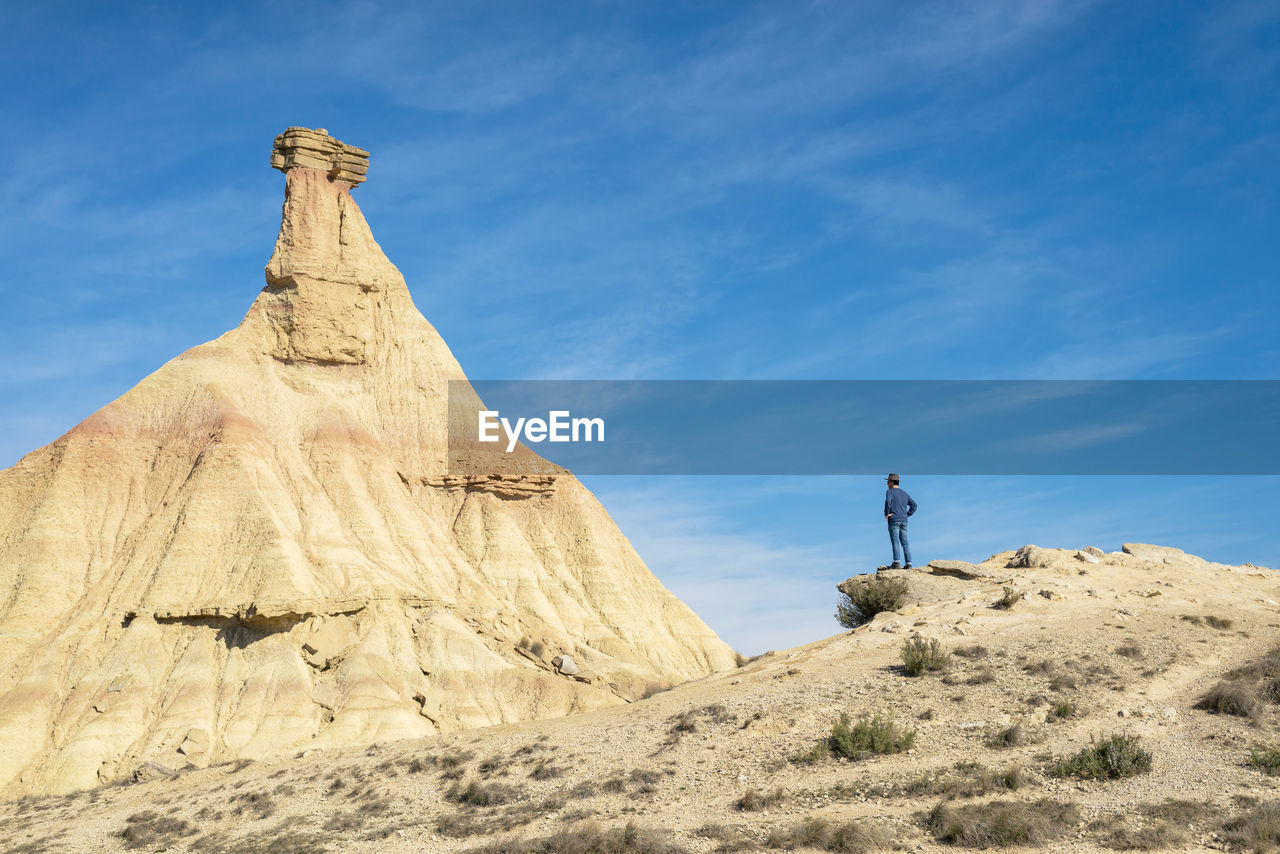 Rear view of a man with hat and sunglasses standing on desert dunes against a rock peak