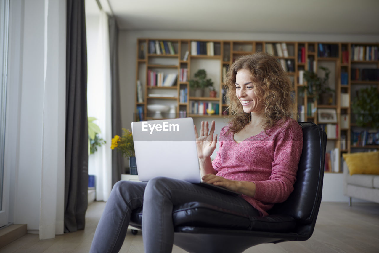 Smiling woman waving hand to video call on laptop while sitting at home