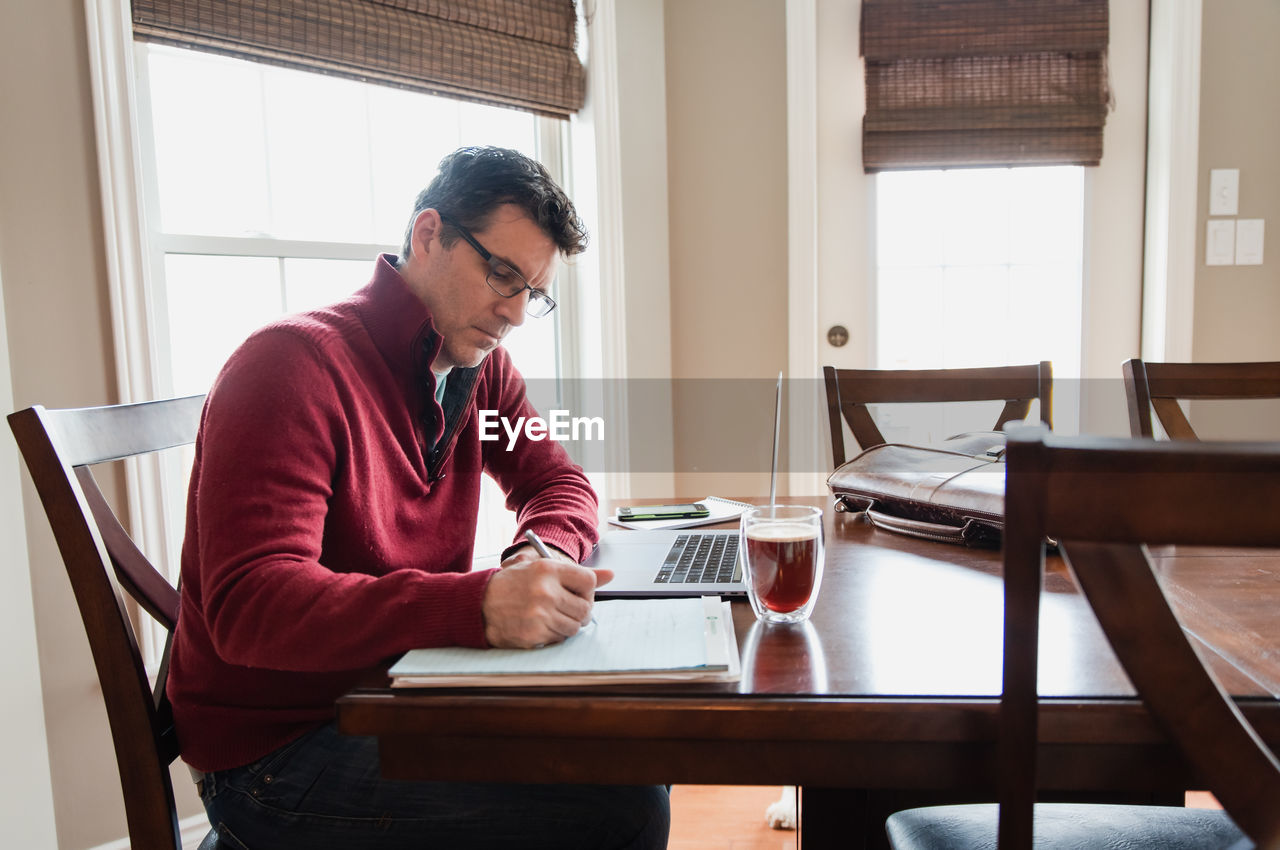 Man in glasses working from home using a computer at a dining table.