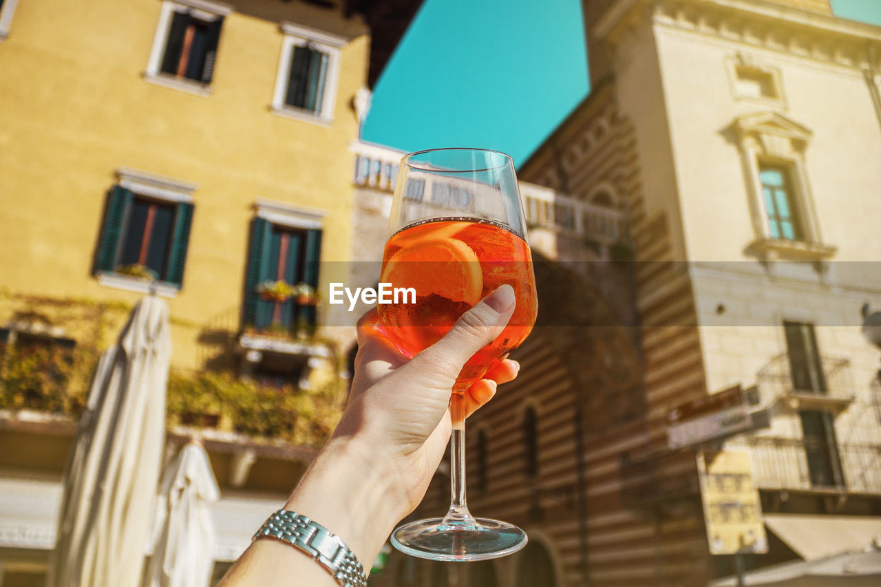 Female hand with glass of orange cocktail spritz near old buildings. sunny day in verona, italy