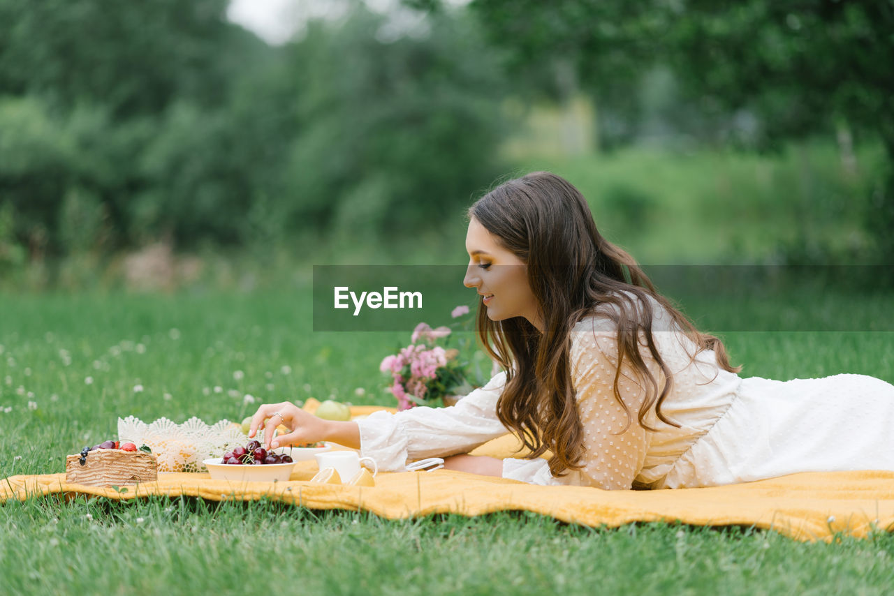 A young beautiful woman in a white dress sits on a blanket on a summer picnic on the green grass