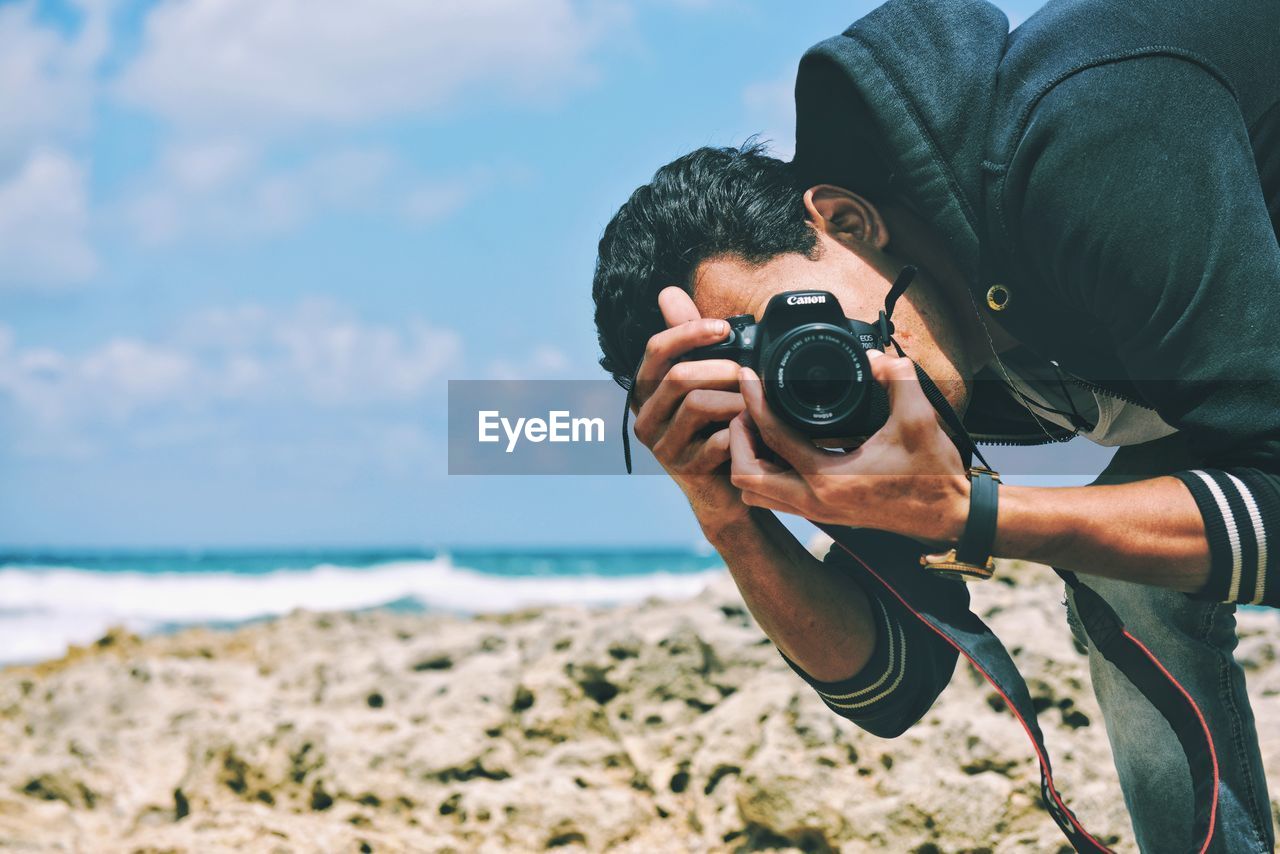 CLOSE-UP OF MAN HOLDING CAMERA WHILE STANDING ON BEACH