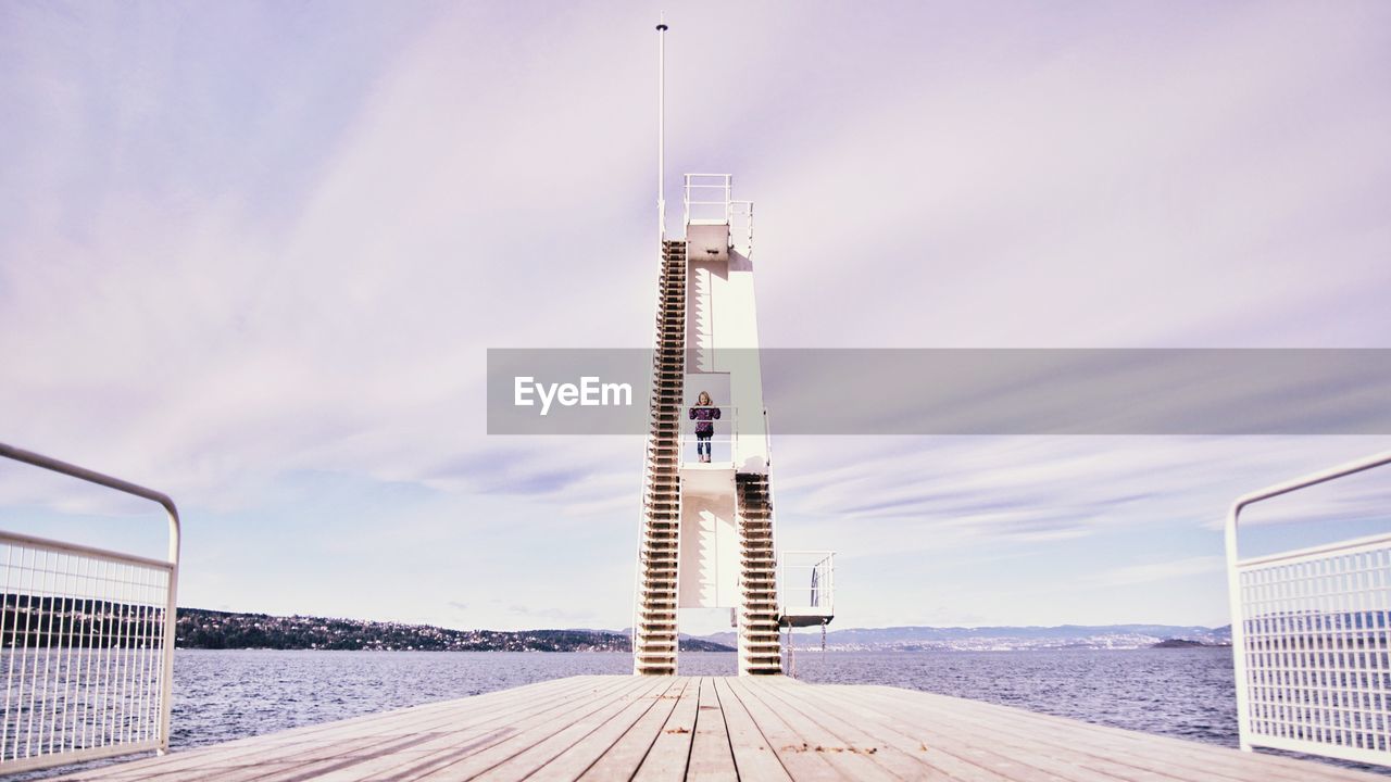 Woman standing on diving platform by river against sky