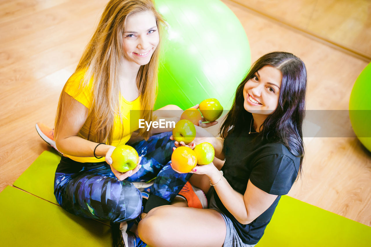 Portrait of smiling young friends holding apples while sitting at yoga studio