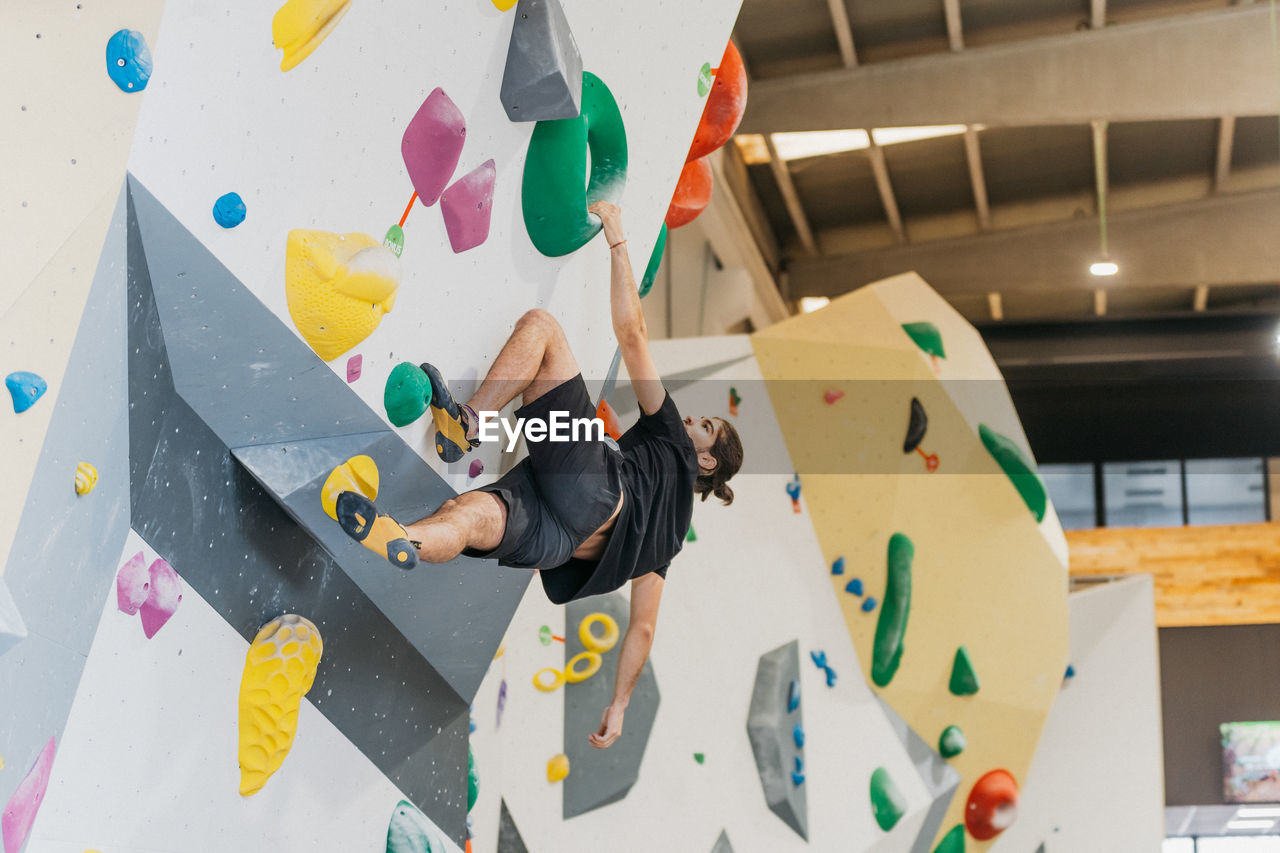 Full body side view of sportive male alpinist climbing wall with colorful grips during intense training in light modern gym