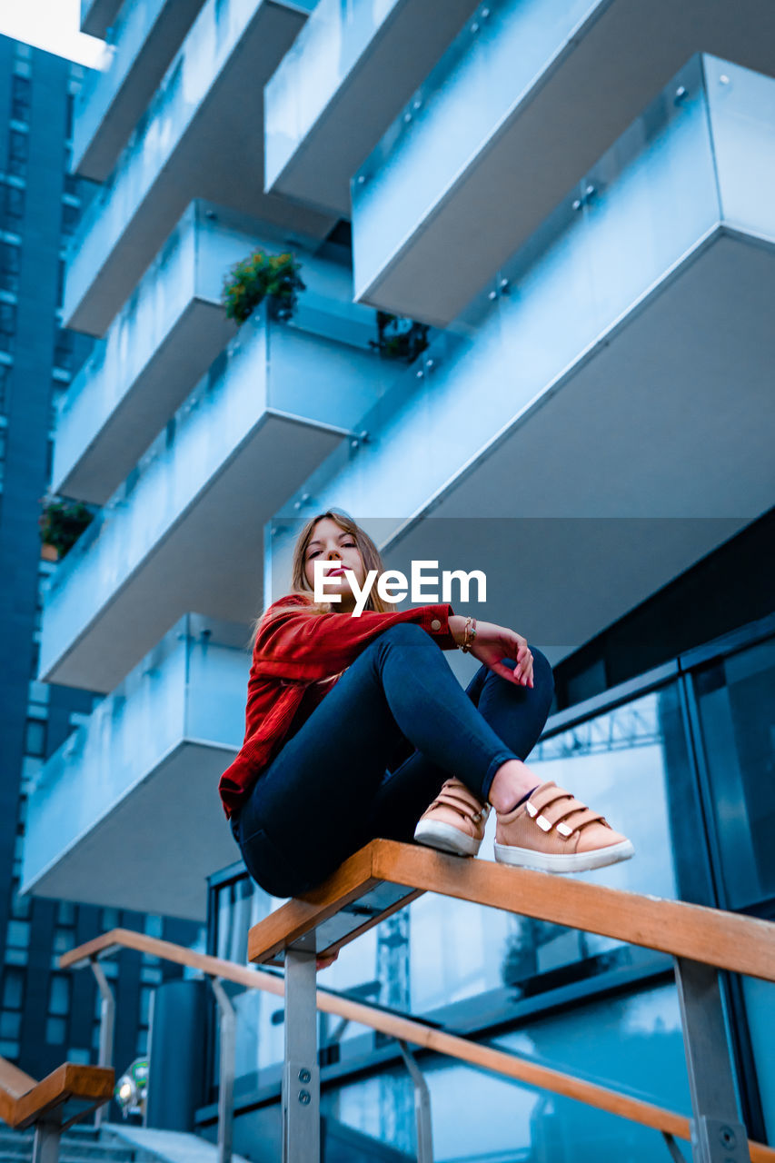 Low angle view of woman sitting on railing against building in city