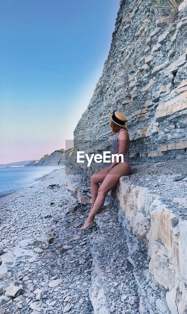 Side view of woman sitting on rocks by sea against sky