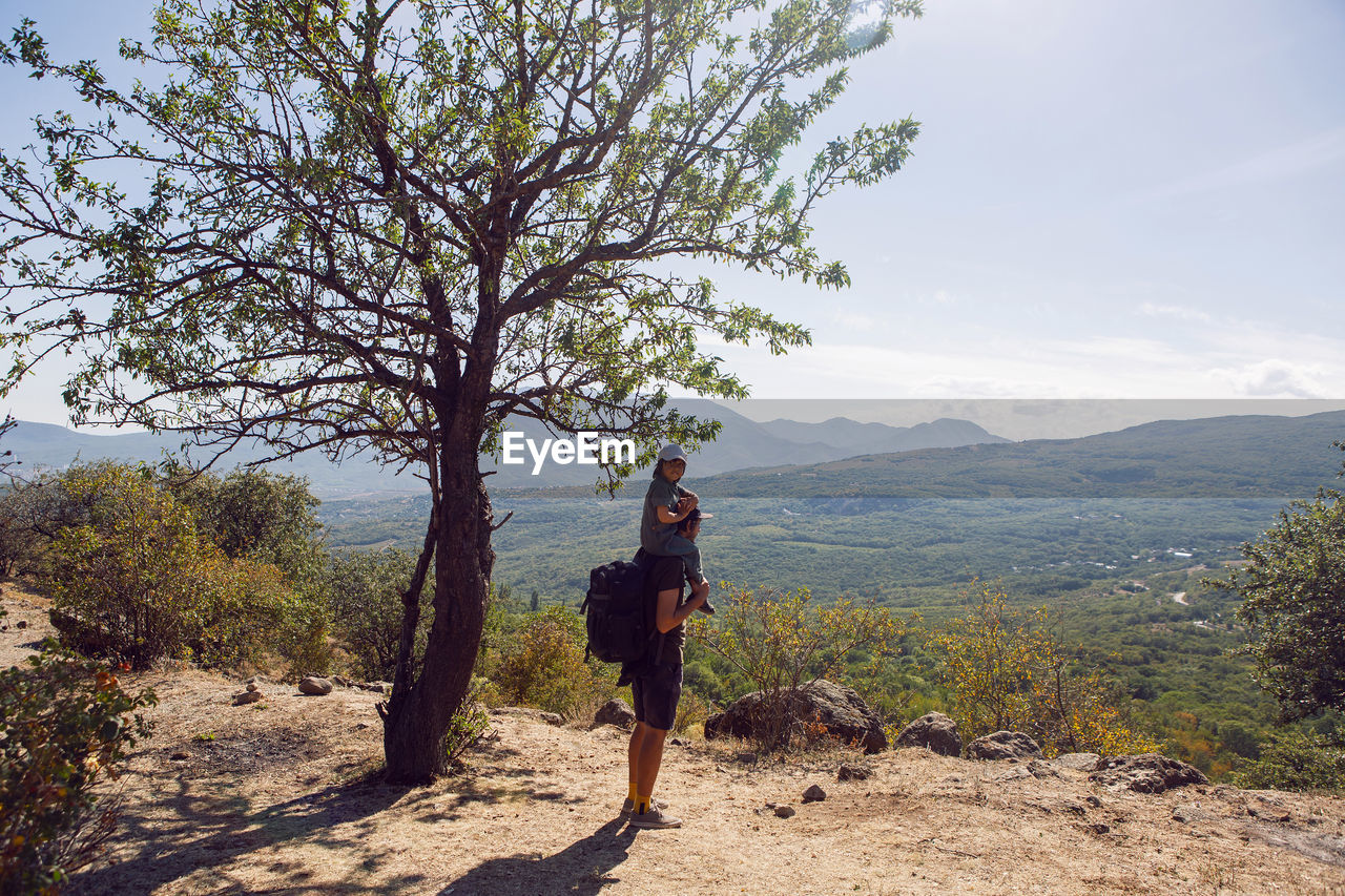 Father with a backpack and his son stands next to a tree on a mountain in the summer in the crimea