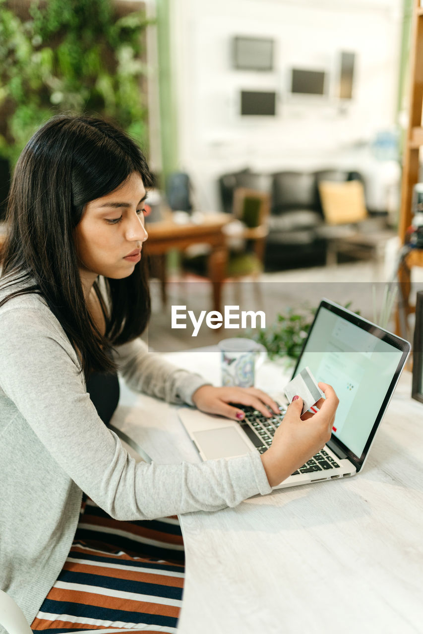 Side view female making purchase with plastic card for order during online shopping via laptop