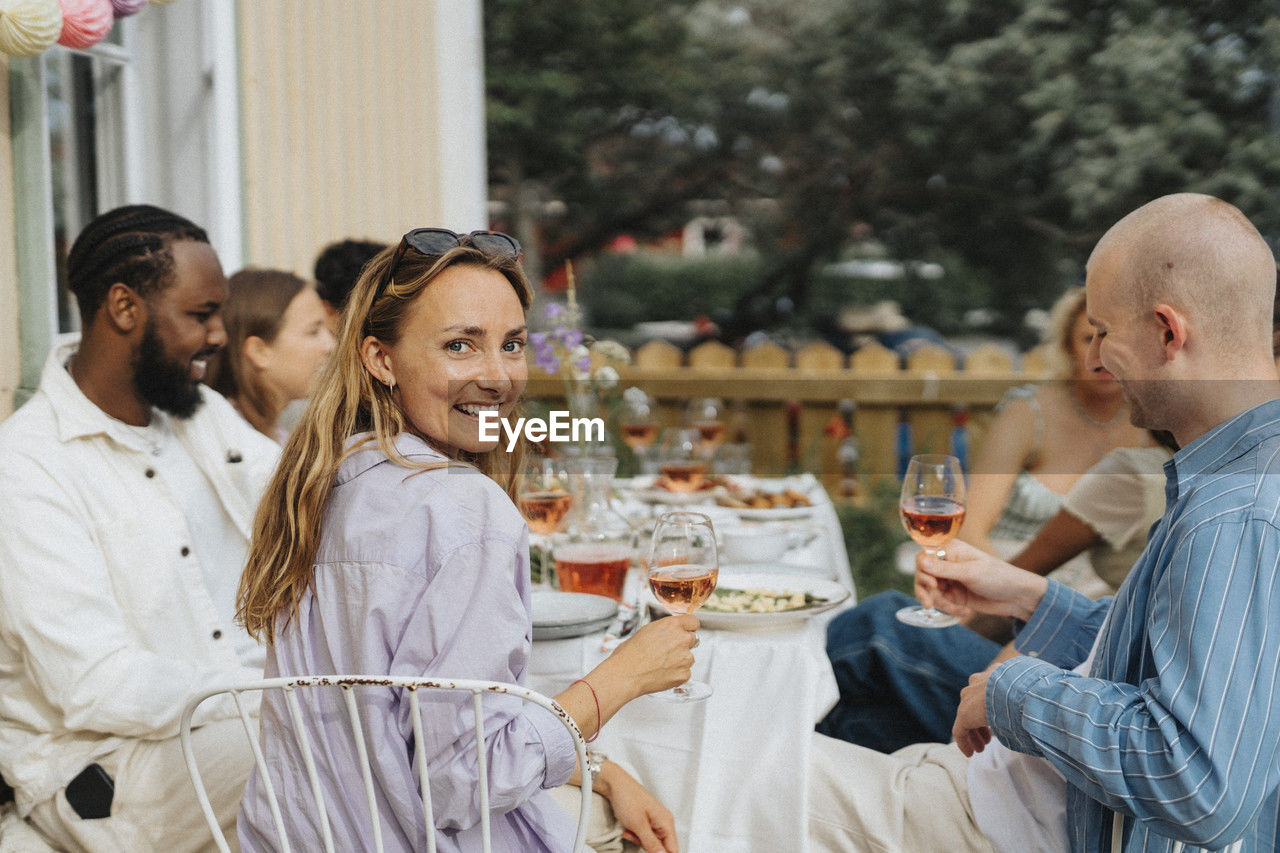 Portrait of smiling young woman looking over shoulder while sitting with friends during dinner party at cafe