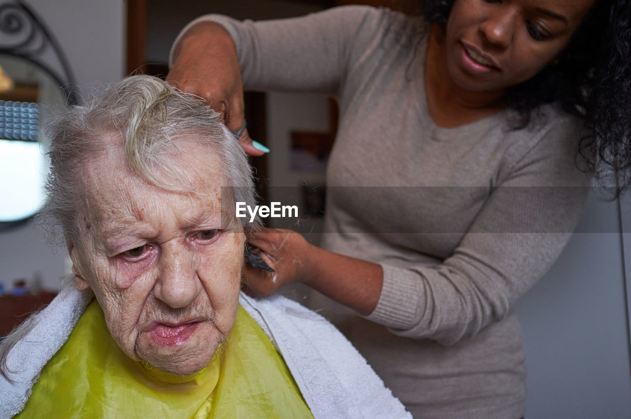 Caring african american woman caregiver, cutting her elderly woman hair at home