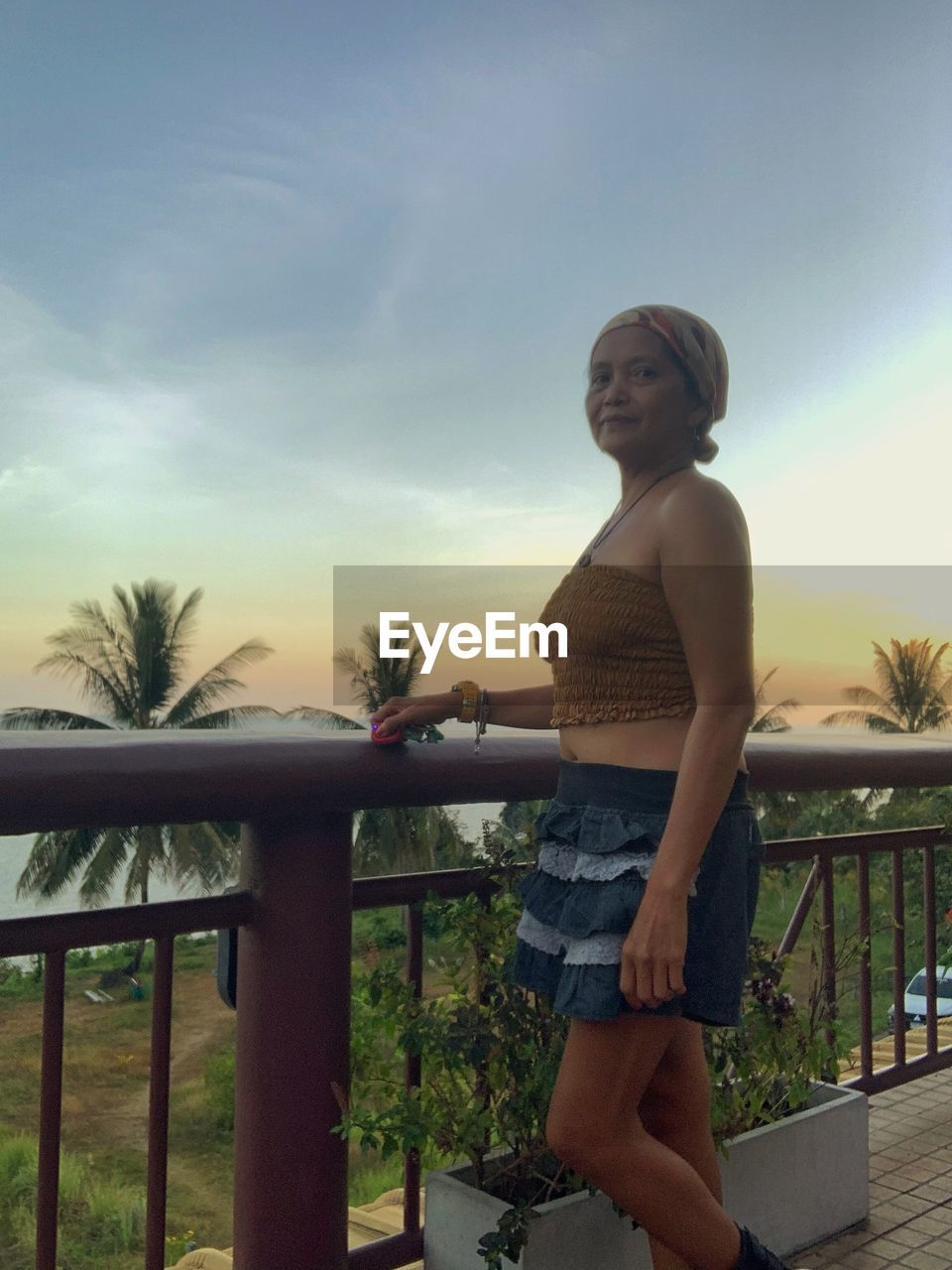 one person, sky, women, adult, vacation, railing, young adult, nature, standing, lifestyles, full length, tropical climate, leisure activity, clothing, casual clothing, palm tree, sunset, architecture, tree, fashion, relaxation, outdoors, trip, looking, holiday, cloud, plant, sitting, hairstyle, limb, human leg, sunlight, smiling, female, day, built structure, happiness, water, beauty in nature, side view, person, shorts, three quarter length, looking away
