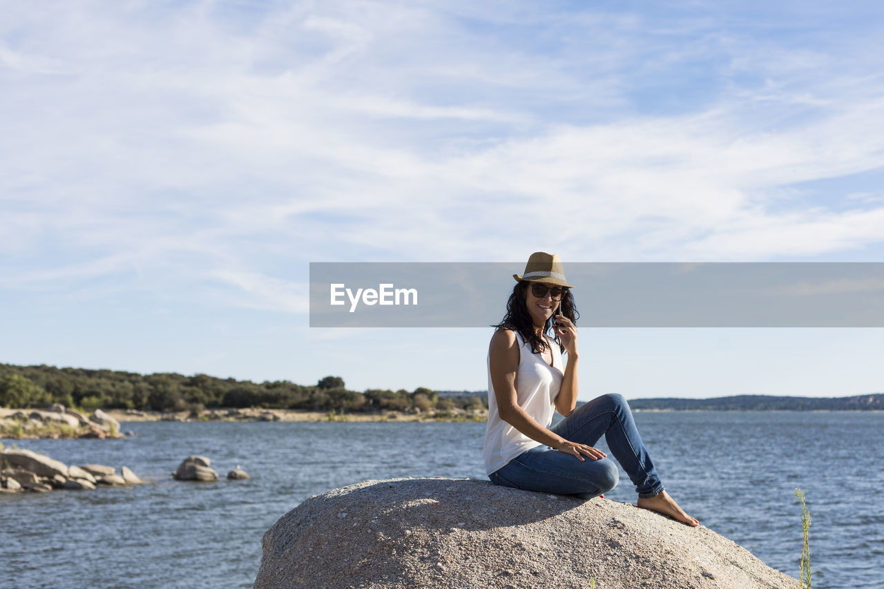 WOMAN SITTING ON ROCK LOOKING AT SEA SHORE AGAINST SKY