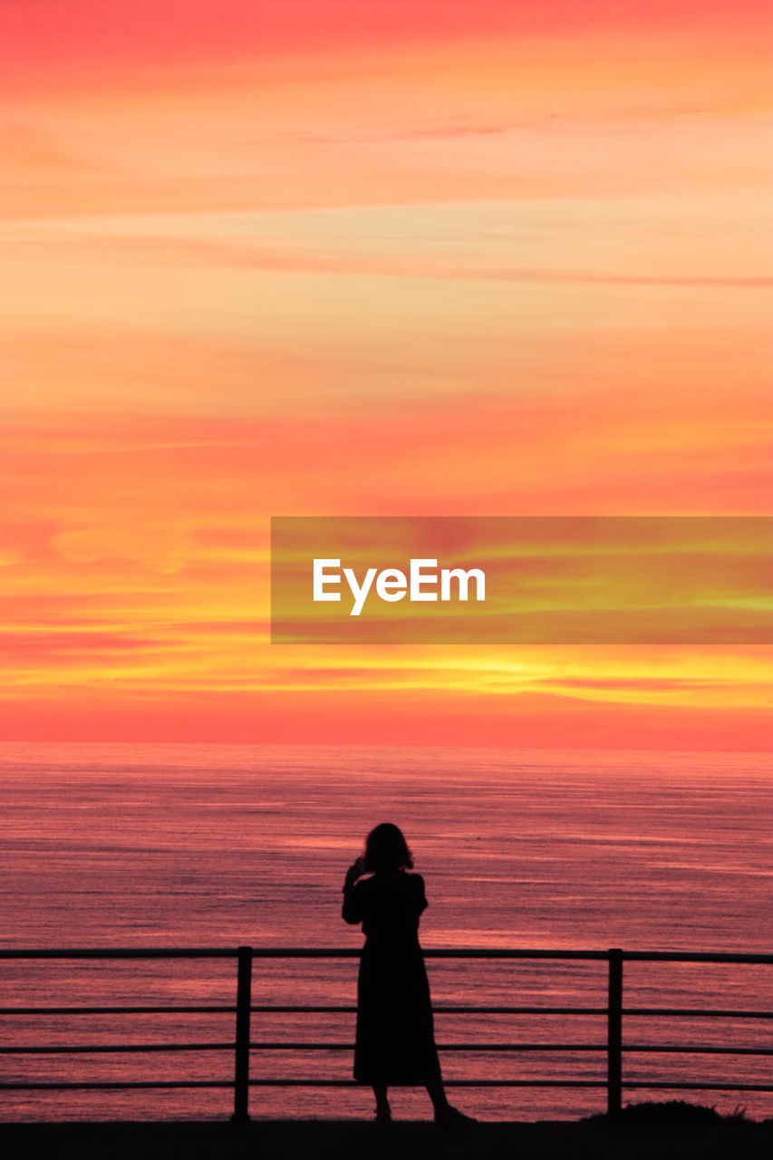 SILHOUETTE WOMAN STANDING ON SEA AGAINST ORANGE SKY