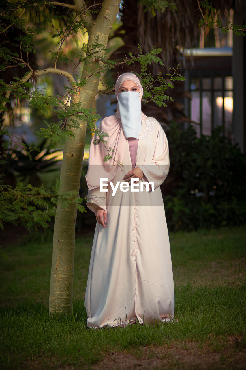 Full length image of veiled woman with white niqab and pink silky dress next to the tree