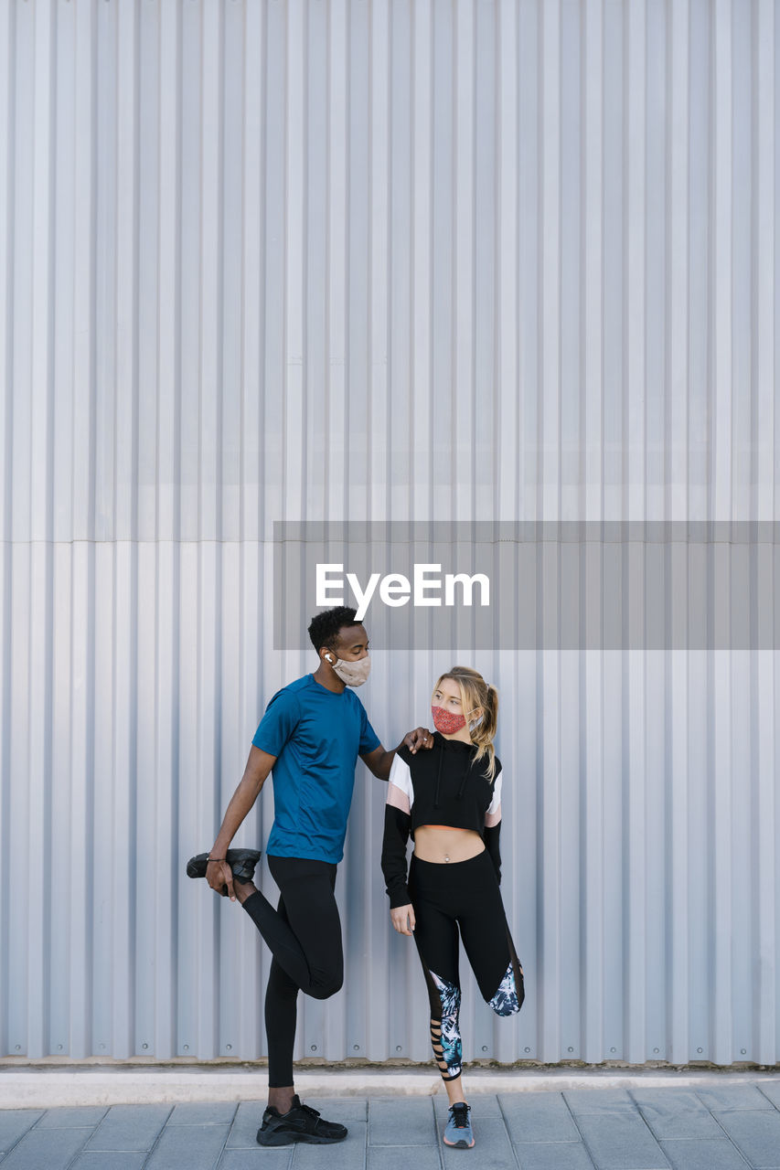 Male and female sportsperson with protective face mask doing warm up exercise while standing against wall during pandemic