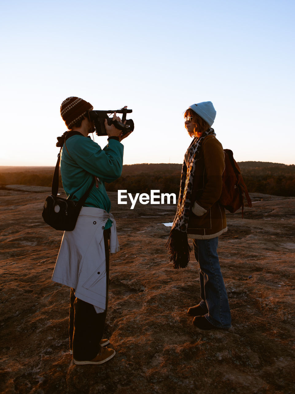 Side view of man photographing woman standing on land against clear sky during sunset