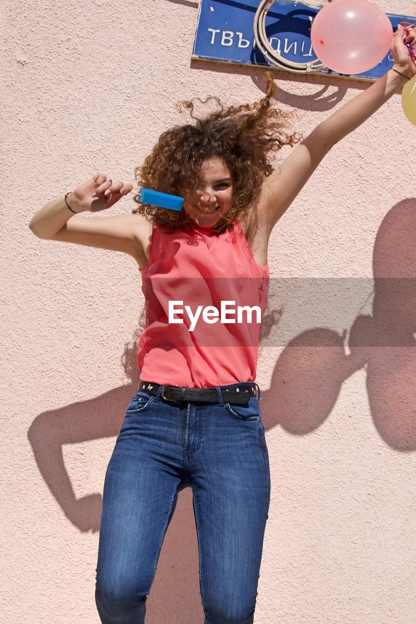 Smiling young woman holding balloons against wall during sunny day