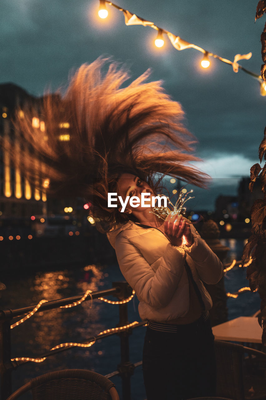 Portrait of smiling young woman tossing hair while holding illuminated lighting equipment at night