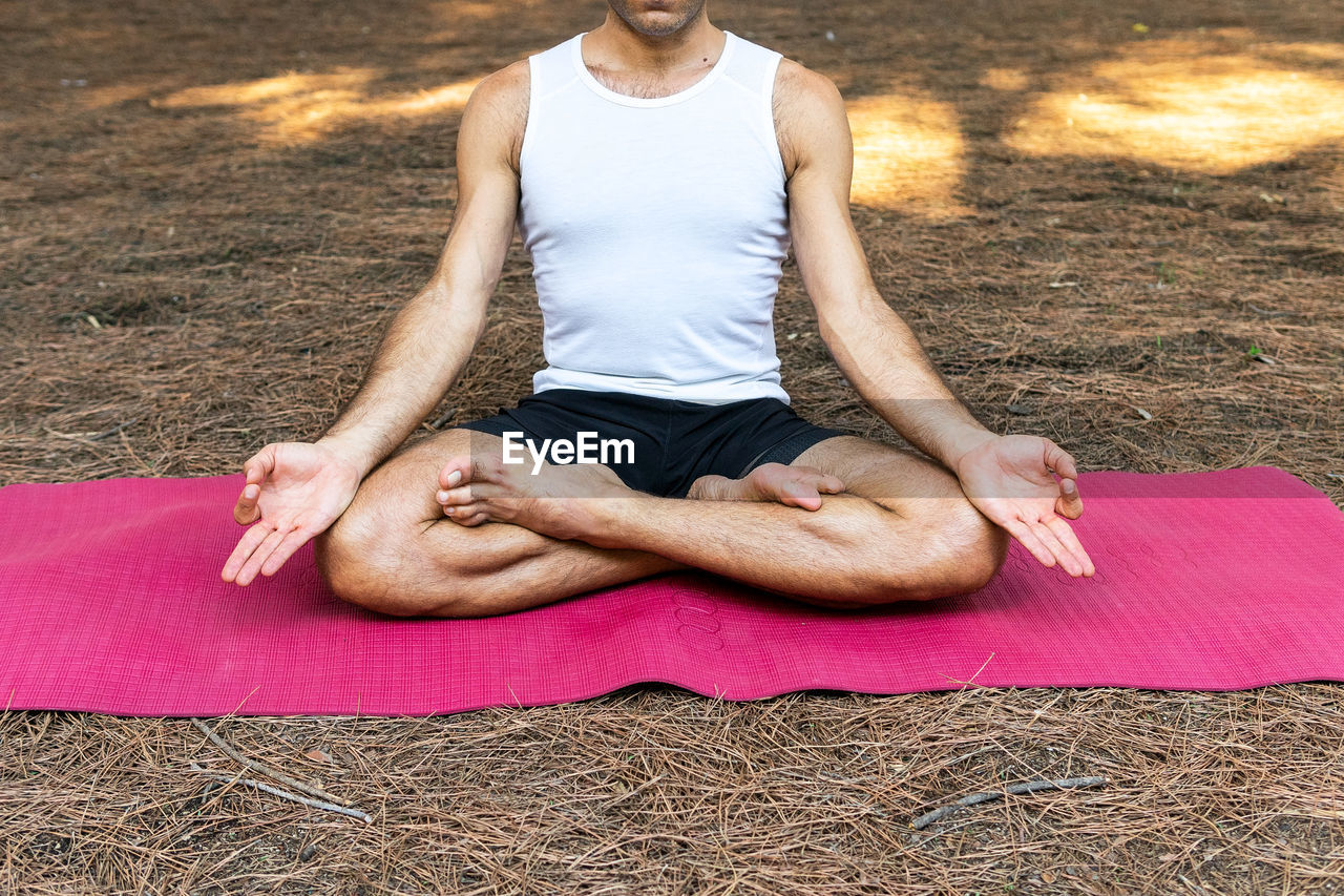 Unrecognizable barefoot male sitting in lotus pose on magenta mat and gesturing mudra during meditation session on sunny summer day in park