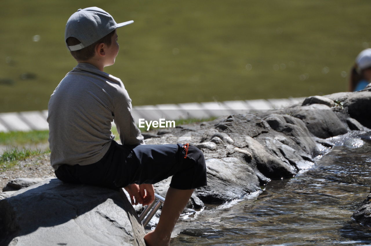 Side view of boy sitting on rock near a stream playing with the water in an outdoor playground