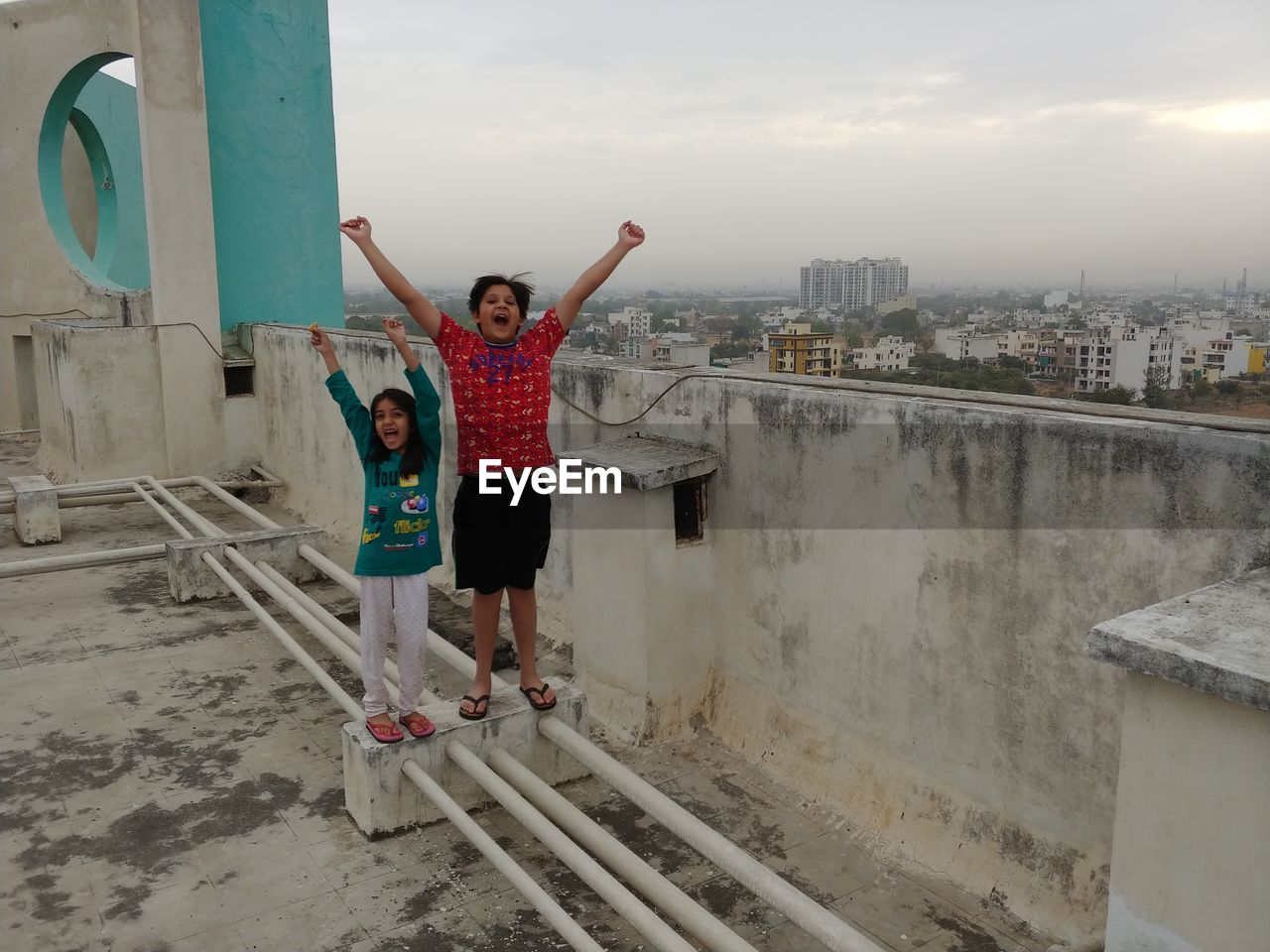 Portrait of happy siblings with arms raised standing on building terrace against cloudy sky