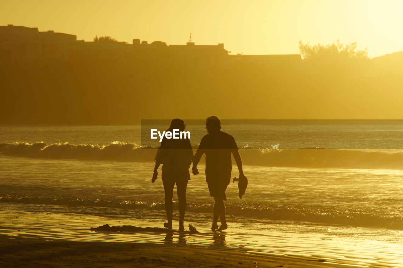 SILHOUETTE FRIENDS STANDING ON SHORE AGAINST SEA DURING SUNSET