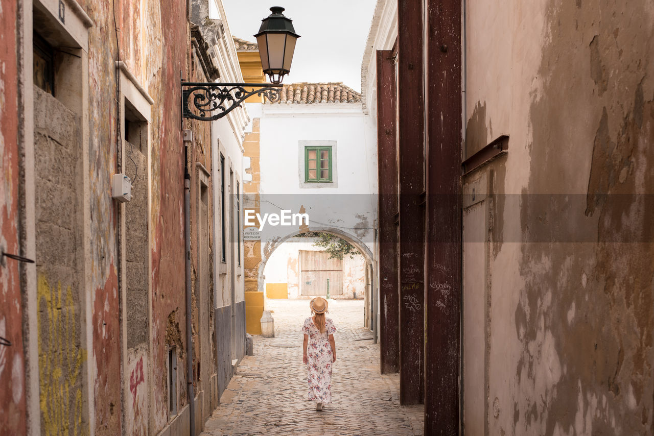 Back view of anonymous young female traveler in casual summer clothes walking along narrow paved street in algarve, portugal