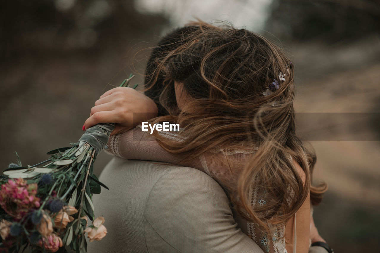 Side view of young guy in suit embracing with lady in wedding dress with bunch of flowers in hand near trees on blurred background