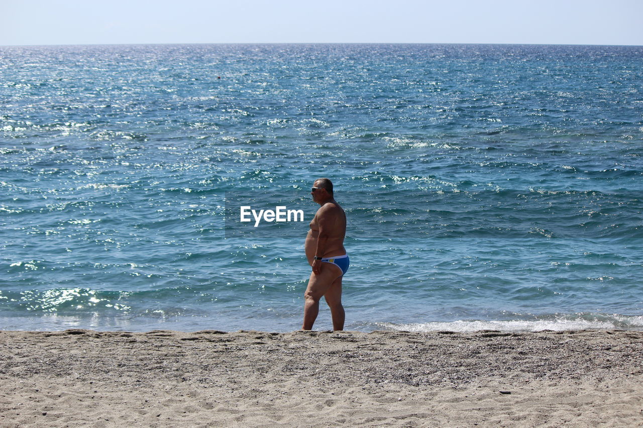 Side view of shirtless overweight man standing on shore at beach during sunny day