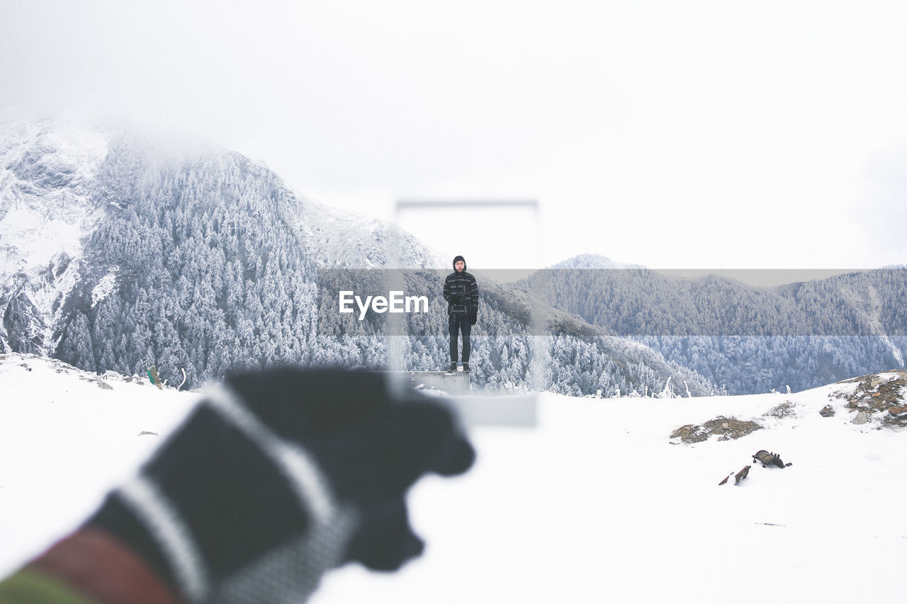 Cropped hand holding frame in front of man standing on snow covered mountain