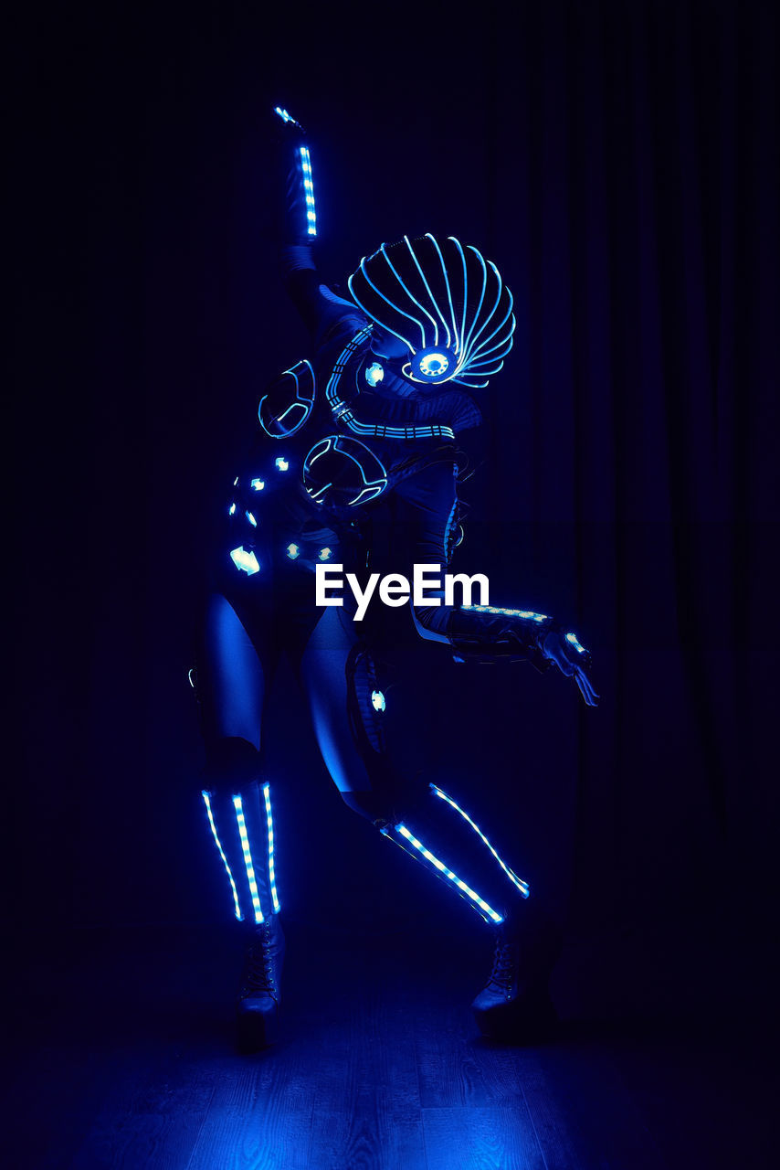 Faceless person in contemporary glowing suit of space cyborg with neon illumination and helmet standing on black background in dark studio person