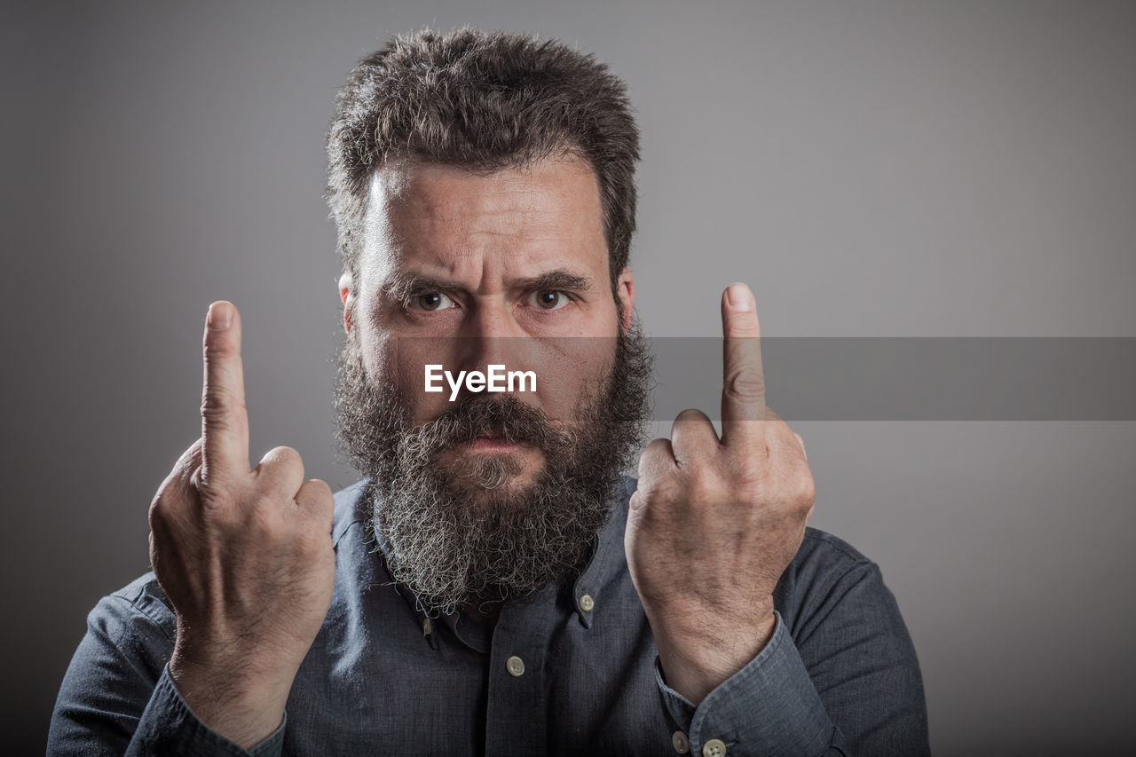 Portrait of bearded man showing middle fingers against gray background