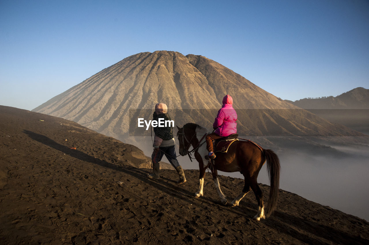 Rear view of person riding horse while man walking on mountain against sky