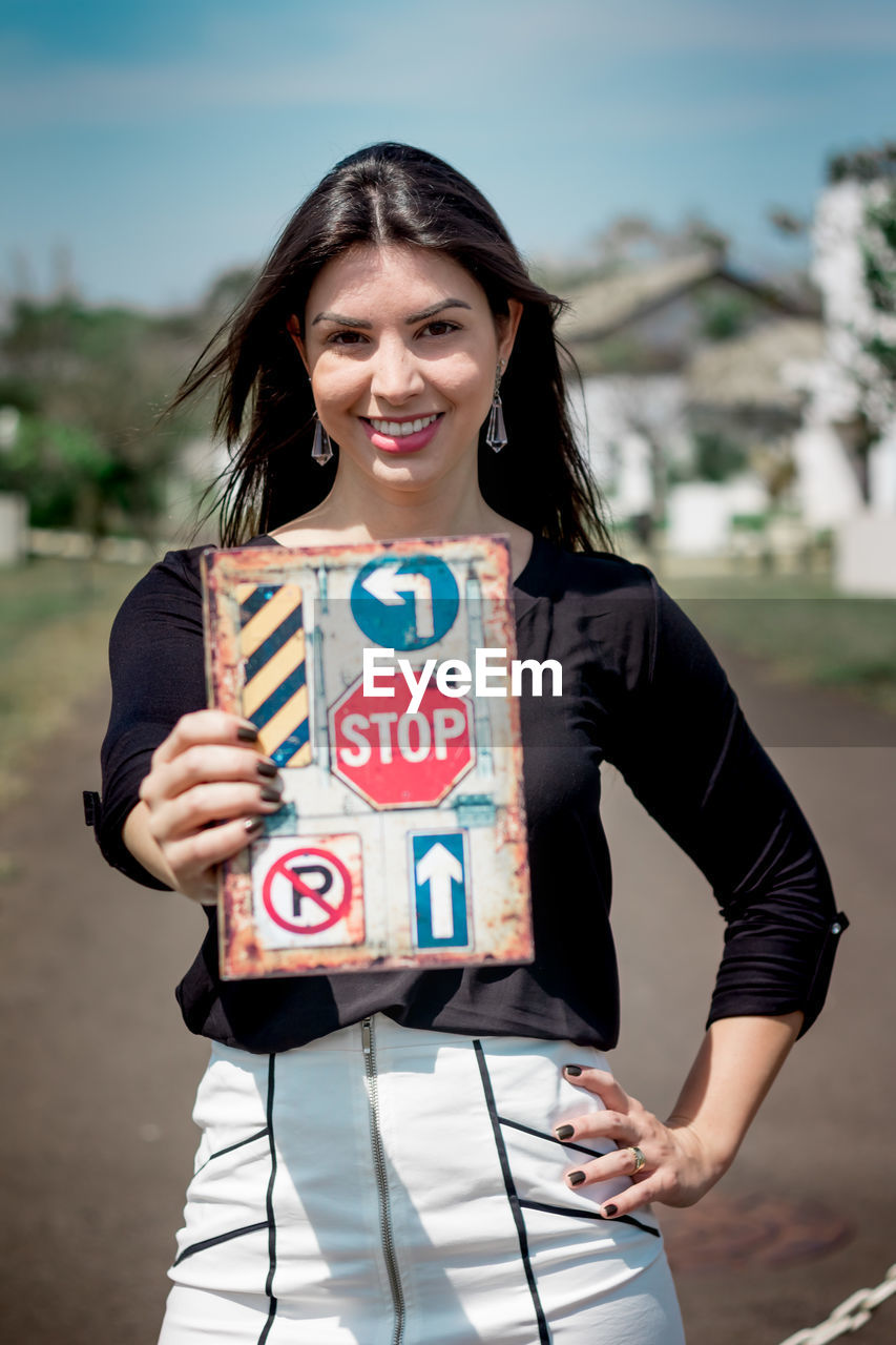 Portrait of smiling young woman holding road signs while standing outdoors