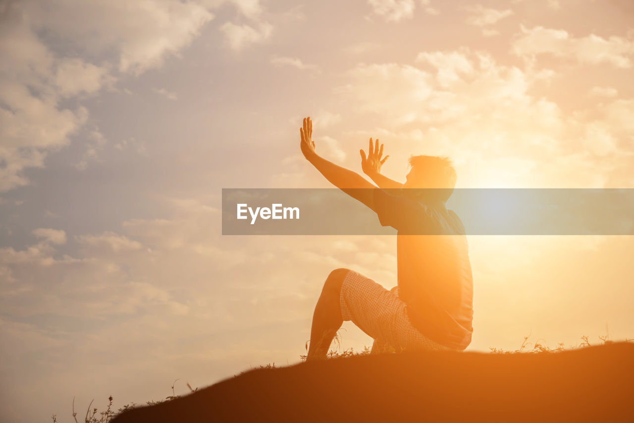 Side view of man gesturing while sitting on land against sky during sunset