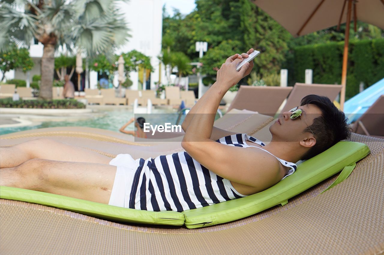 Mid adult man using phone while lying on lounge chair at poolside
