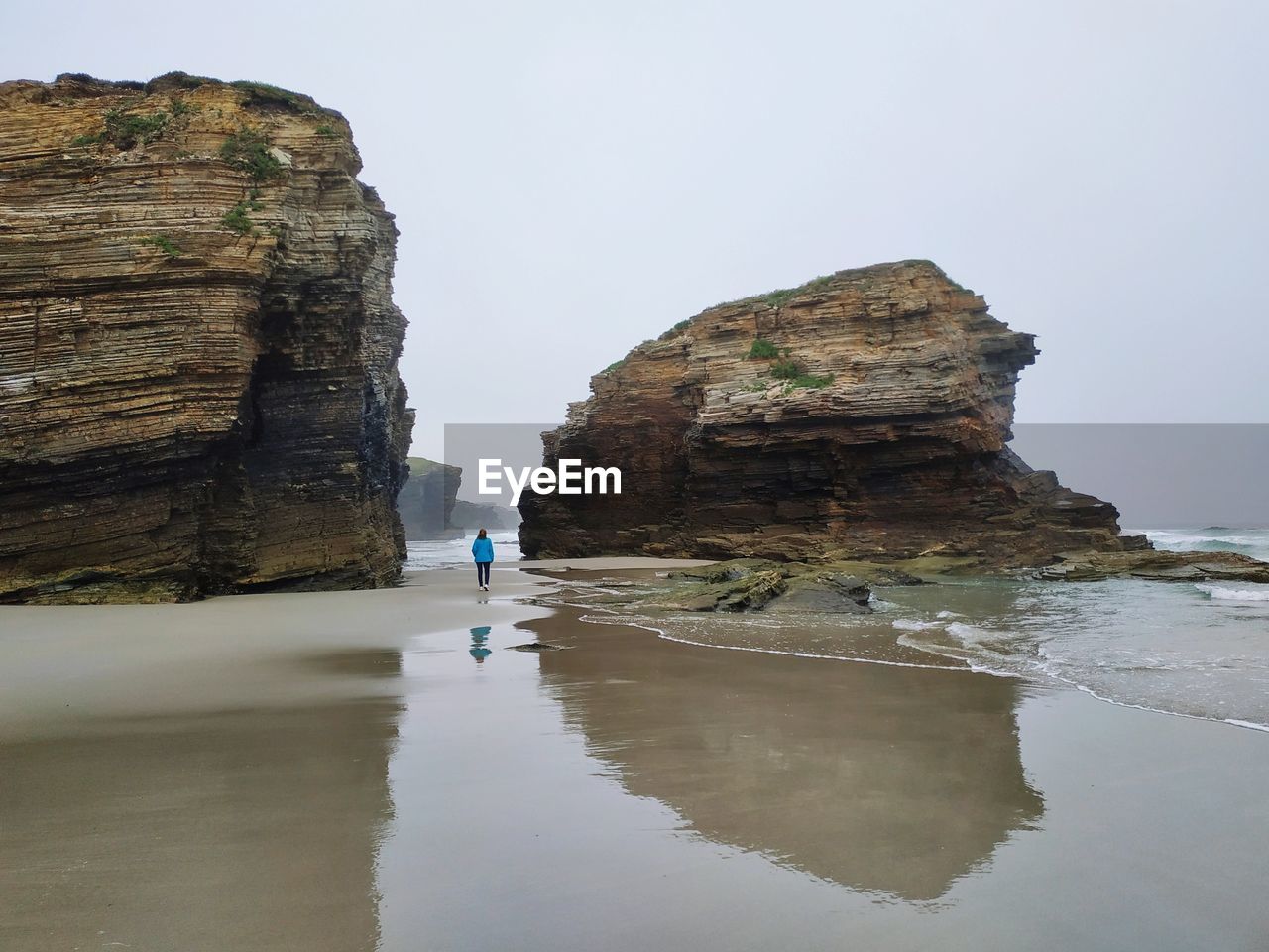 Distant view of woman walking at beach by rock formations against sky