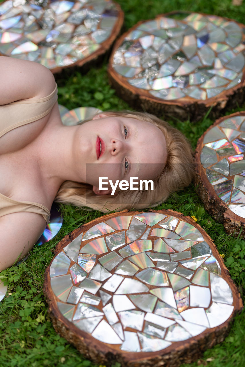 Top view portrait of a young woman lying on a lawn among stumps covered with a mirror