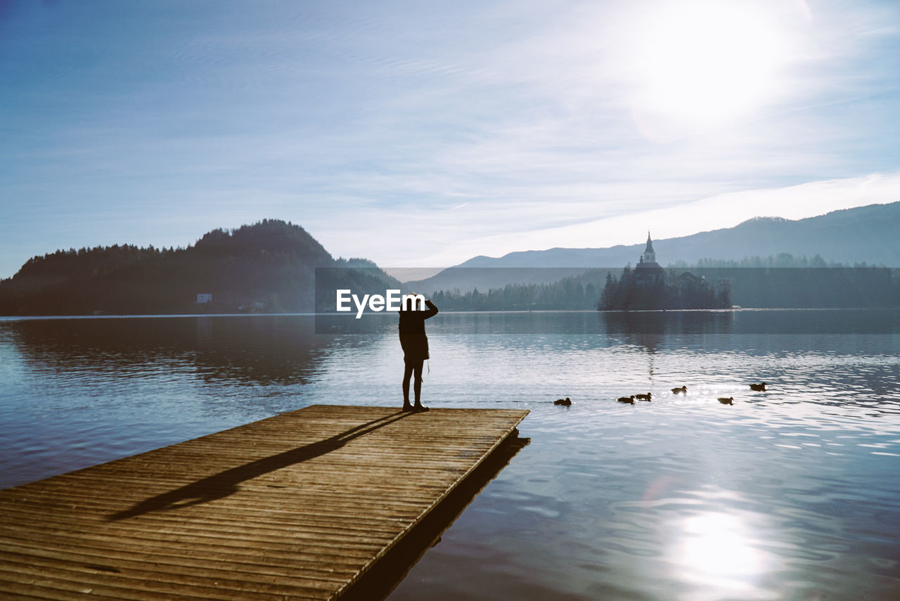 Woman standing on pier at lake against sky during sunset