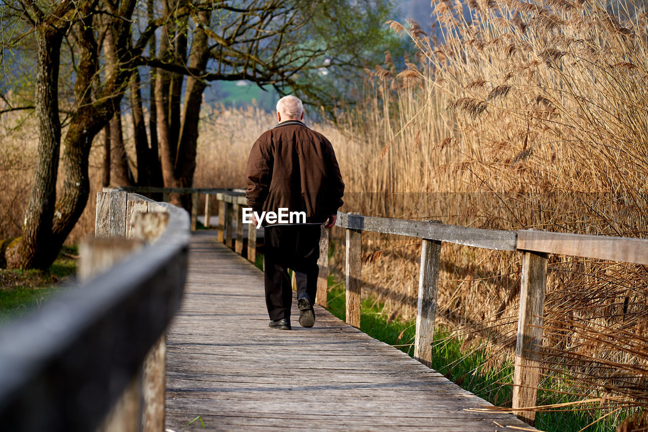 REAR VIEW OF MAN WALKING ON FOOTPATH BY FOOTBRIDGE