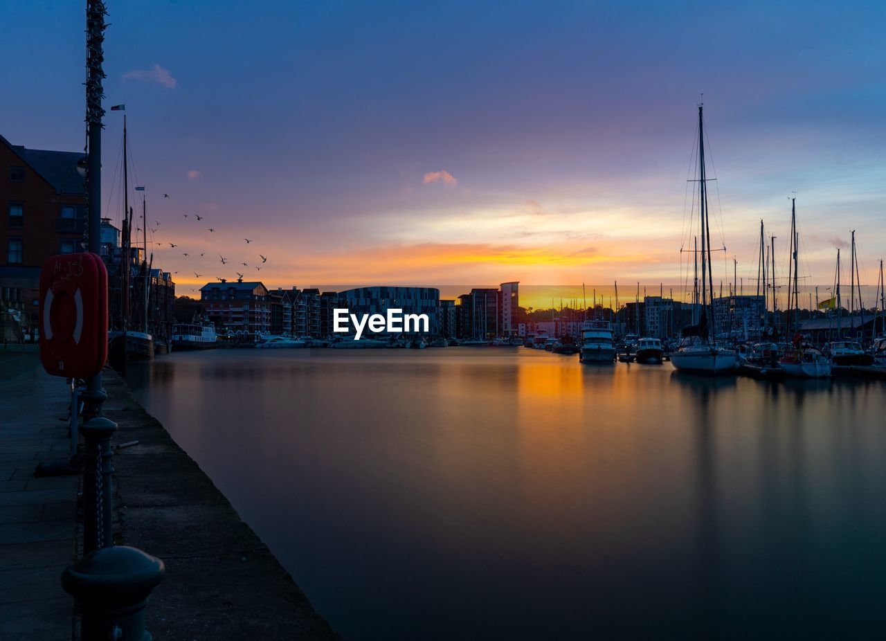 Sailboats moored on harbor by buildings against sky at sunset