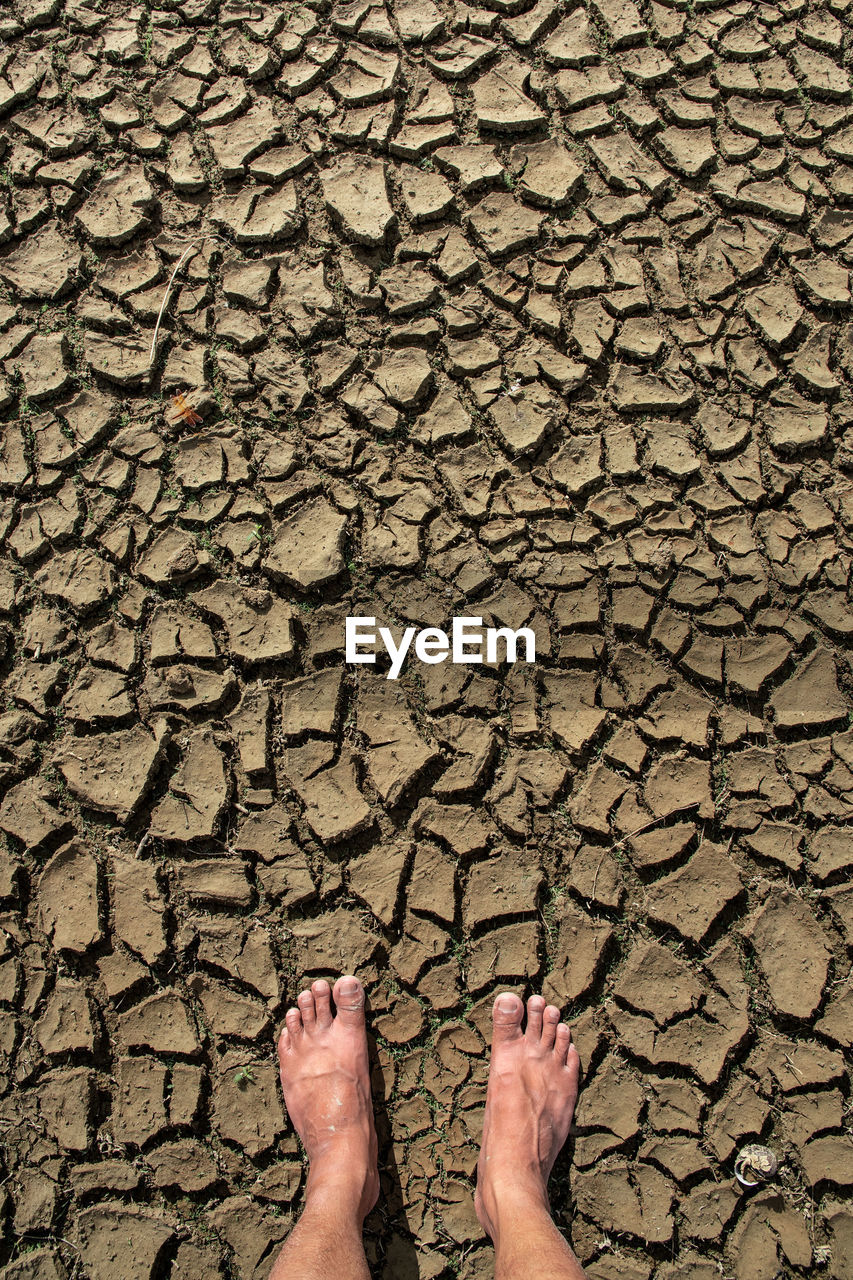 soil, drought, cracked, one person, low section, climate, nature, human leg, dry, land, arid climate, high angle view, standing, personal perspective, environment, day, lifestyles, barefoot, road surface, dirt, pattern, outdoors, scenics - nature, human foot, leisure activity, environmental issues, directly above, barren, wall, limb, human limb, textured, asphalt, mud, sand, field, adult, brown, sunlight