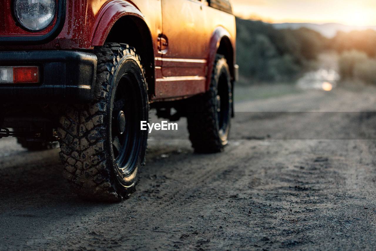 Cropped modern red off road car parked on dusty dirt road leading through forest in summer evening in countryside