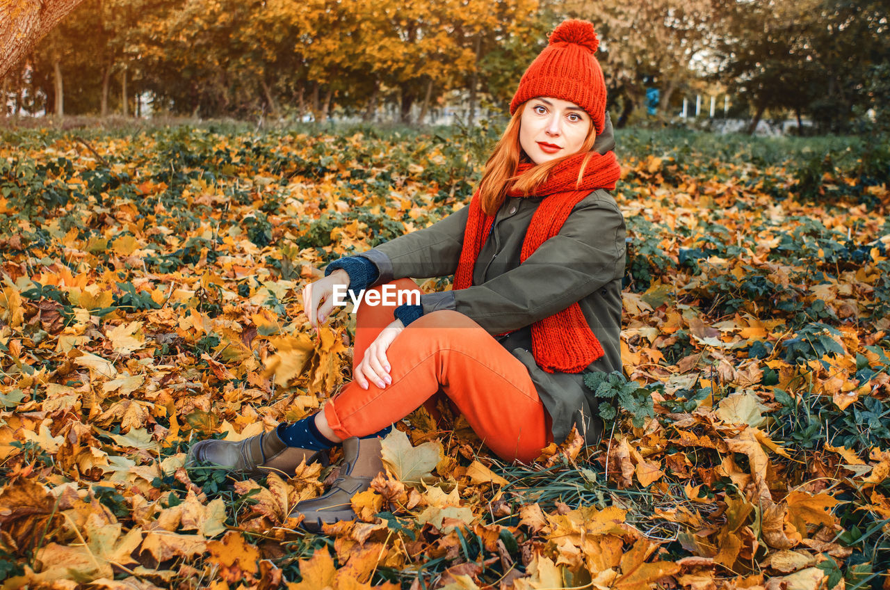 Autumn portrait of cute girl in orange warm knitted hat and orange scarf sitting in fallen leaf