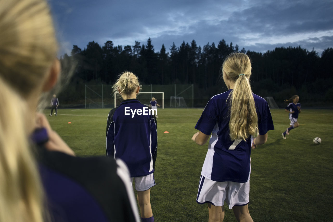 Rear view of girls standing on soccer field against sky at dusk