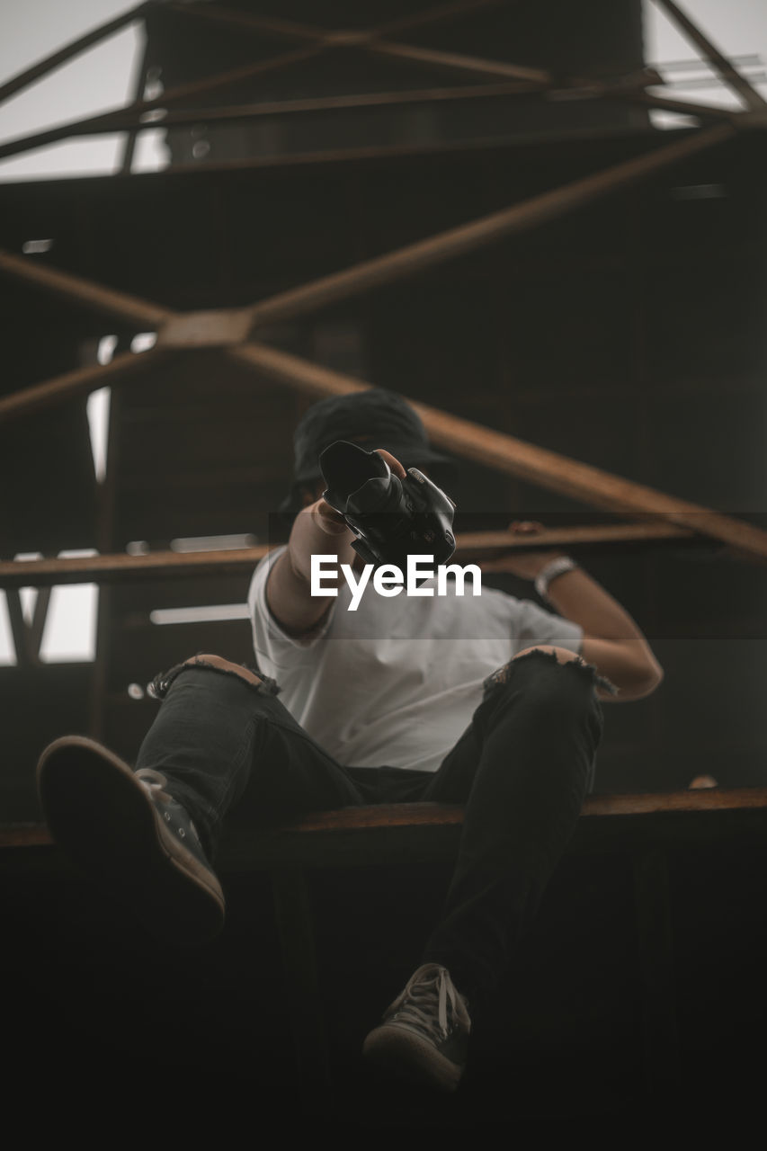 Low angle view of man holding camera while sitting against railing