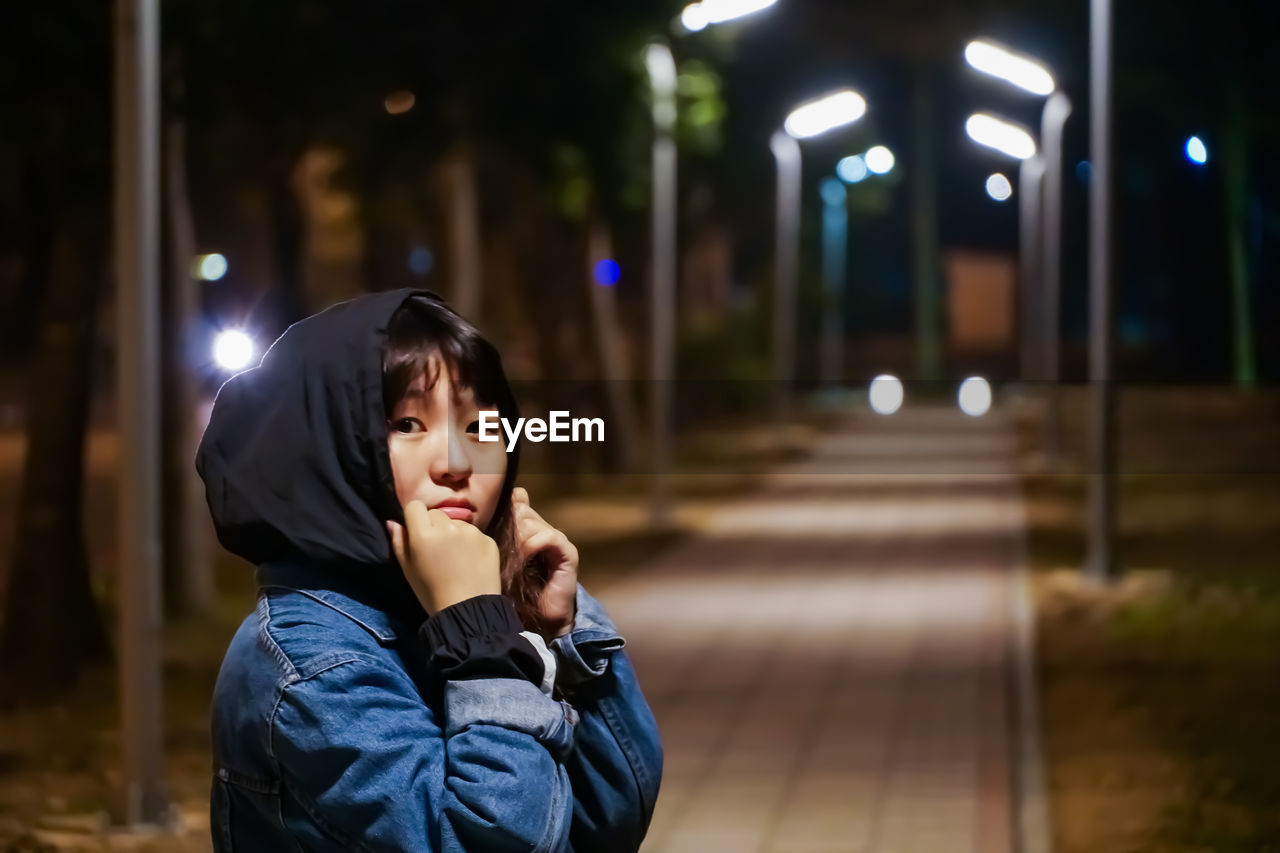 Portrait of woman standing on footpath against illuminated lights at night